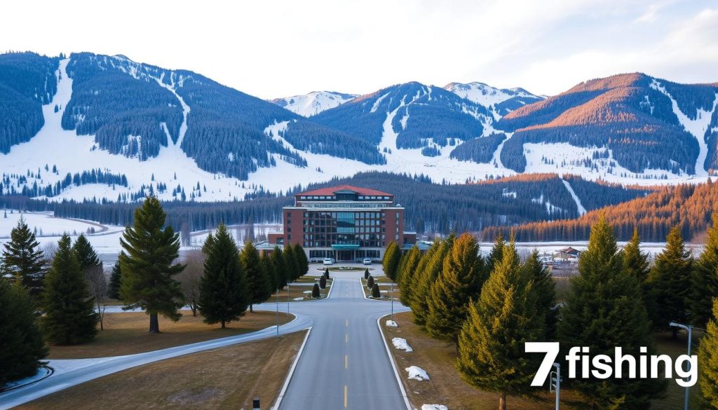A picturesque landscape of the Petros Hotel nestled in the heart of the Bukovel ski resort. The hotel stands tall amidst the snow-capped peaks, its modern architecture blending seamlessly with the natural surroundings. In the foreground, a paved road leads to the hotel's entrance, flanked by neatly groomed trees and well-maintained pathways. The middle ground features the hotel's expansive grounds, with ski slopes and lifts visible in the distance. The background is dominated by the majestic Carpathian mountains, their slopes dotted with lush evergreen forests. The scene is bathed in a warm, golden light, creating a cozy and inviting atmosphere. In the bottom right corner, the "7fishing" brand logo adds a touch of branding to the image.
