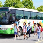 A vibrant scene depicting a colorful bus designed for transporting children to a summer camp, parked outside a picturesque camp setting. In the foreground, smiling children, aged 7 to 12, are ready to board the bus, wearing casual, comfortable clothing, and carrying backpacks filled with camp gear. The middle ground features the bus prominently, with cheerful camp-themed decorations, while staff members wearing matching uniforms assist the children with boarding. In the background, lush green trees and a bright blue sky set a welcoming atmosphere, with the camp building peeking through the foliage. The lighting is bright and sunny, creating a joyful and exciting mood, captured from a slightly elevated angle to emphasize the dynamic interaction between the children and the camp environment.